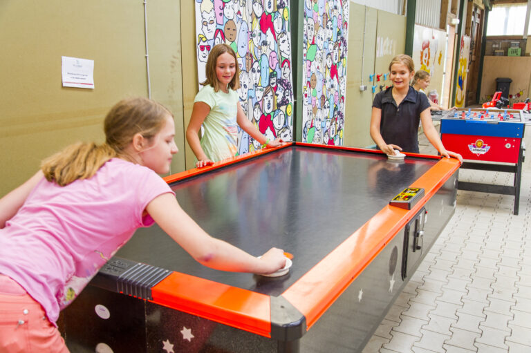 Drei Mädchen spielen in der TeensLounge Airhockey, während im Hintergrund ein Kicker steht und bunte Wandbilder die Halle bei Gut Landegge beleben.