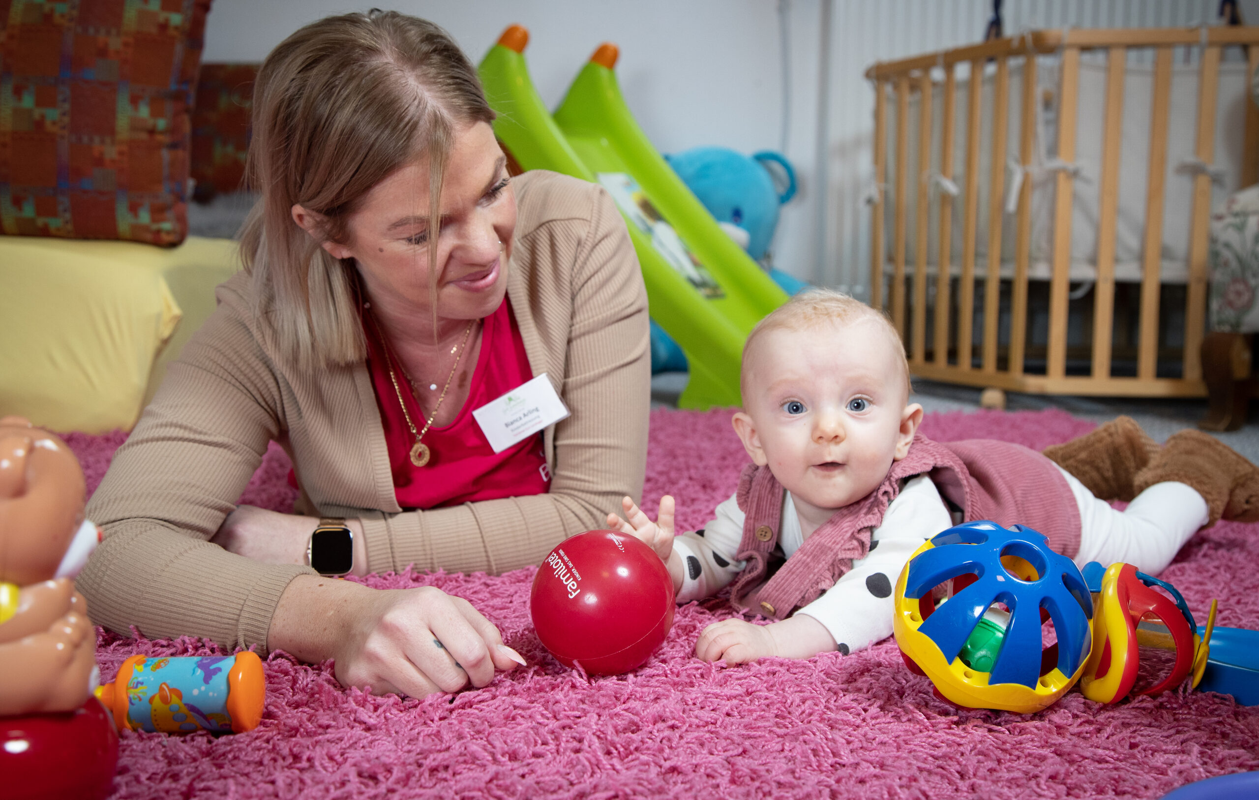 Eine Betreuerin schaut lächelnd auf ein Baby, das auf einer Spielmatte zwischen buntem Greifspielzeug und rotem Ball liegt.