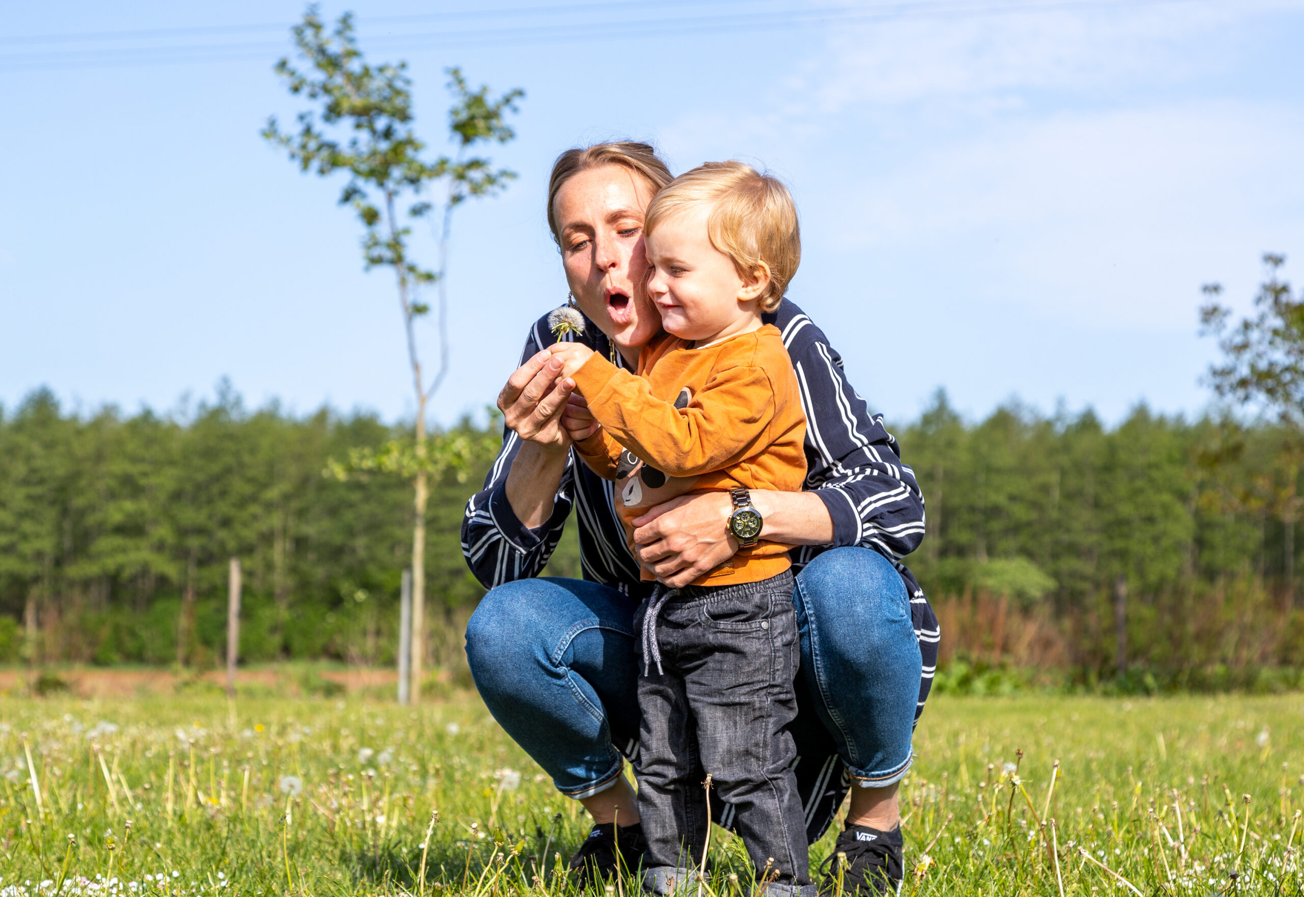 Eine Mutter und ihr Kleinkind betrachten gemeinsam eine Pusteblume auf einer sonnigen Wiese mit jungen Bäumen im Hintergrund.