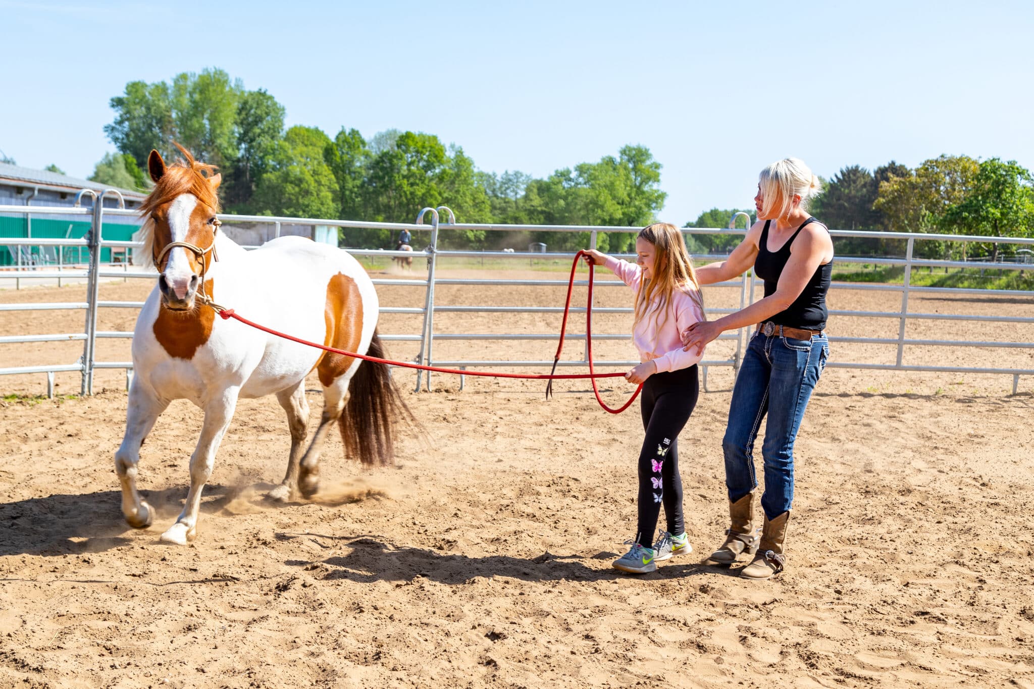 Auf dem Reitplatz führt ein Mädchen ein braun-weißes Pony am roten Seil; eine Erwachsene begleitet sie beim Familienurlaub auf Gut Landegge.