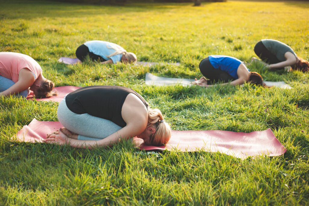 Mehrere Frauen üben gemeinsam Yoga auf Matten im Gras und beugen sich auf einer sonnigen Wiese in die Kindeshaltung.