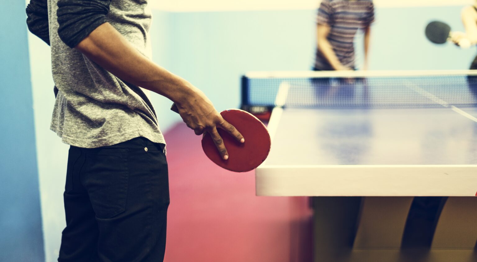 In der Fitness- und Freizeitumgebung hält eine Person einen roten Tischtennisschläger am Tisch, während im Hintergrund weitere Spieler zu sehen sind.