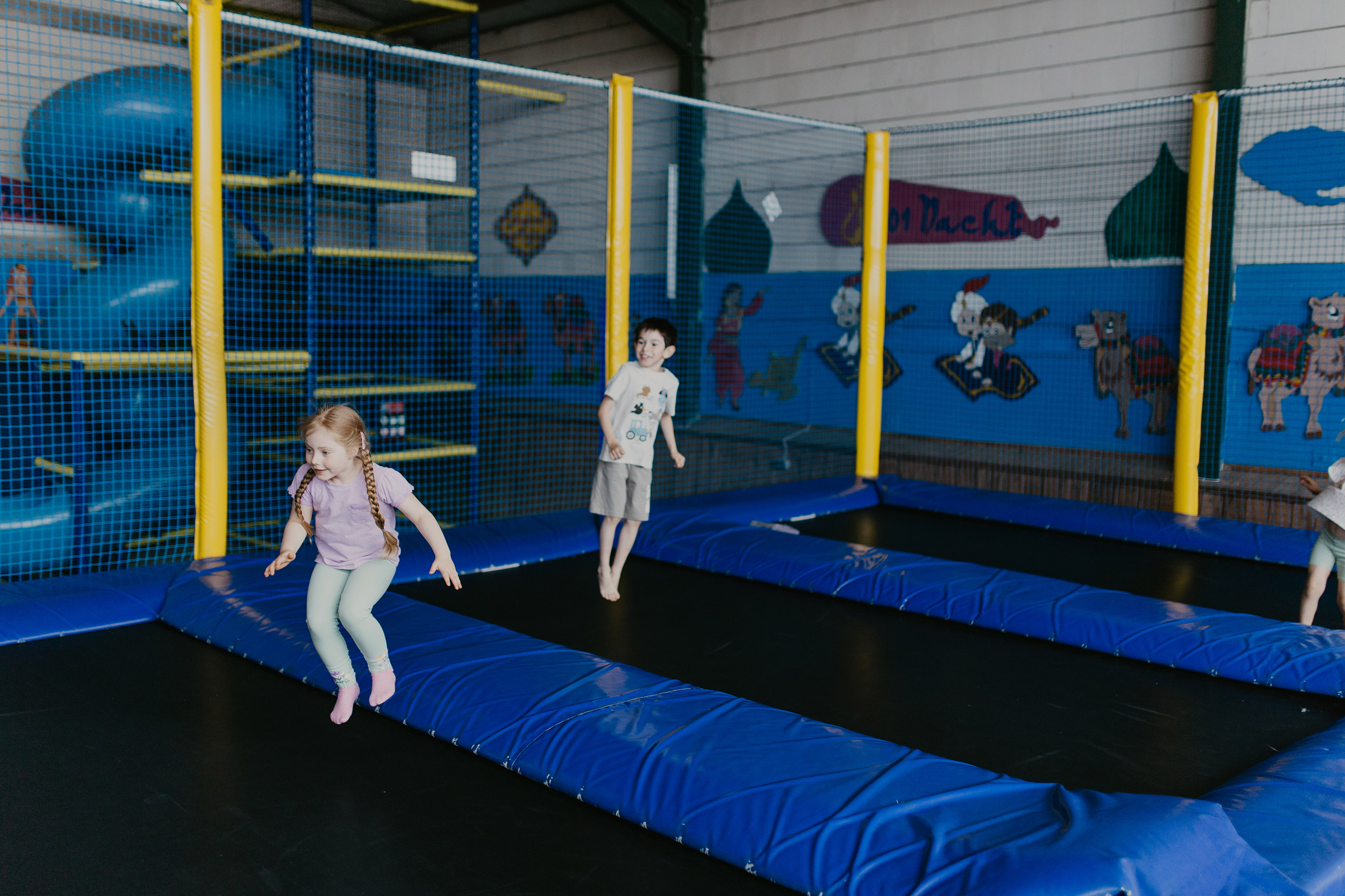 Kinder springen auf Trampolinbahnen in einer Indoor-Spielhalle; Sicherheitsnetze und bunte Wandbilder umgeben die Anlage.