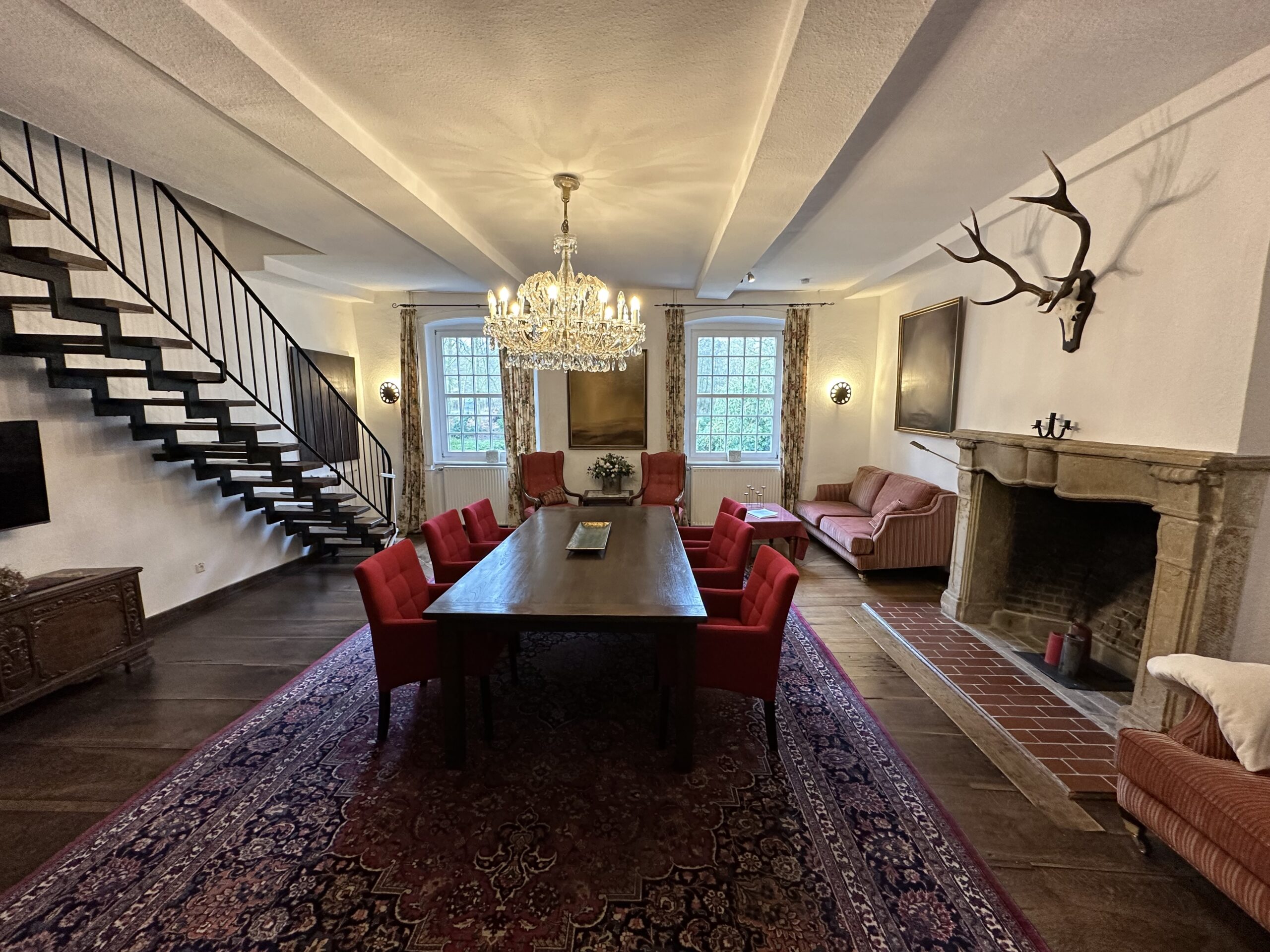 Ein eleganter Saal mit langem Esstisch, roten Polsterstühlen, offener Treppe und großem steinernem Kamin unter einem Kronleuchter.