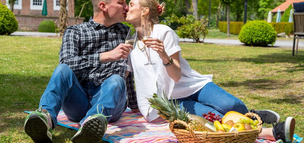 Familienhotel Gut Landegge im Emsland: Eltern machen ein romantisches Picknick auf dem Gut. Sie stoßen mit Sekt an und geben sich einen Kuss. Ein leckerer Korb voller Früchte steht vor ihnen auf der Picknickdecke.