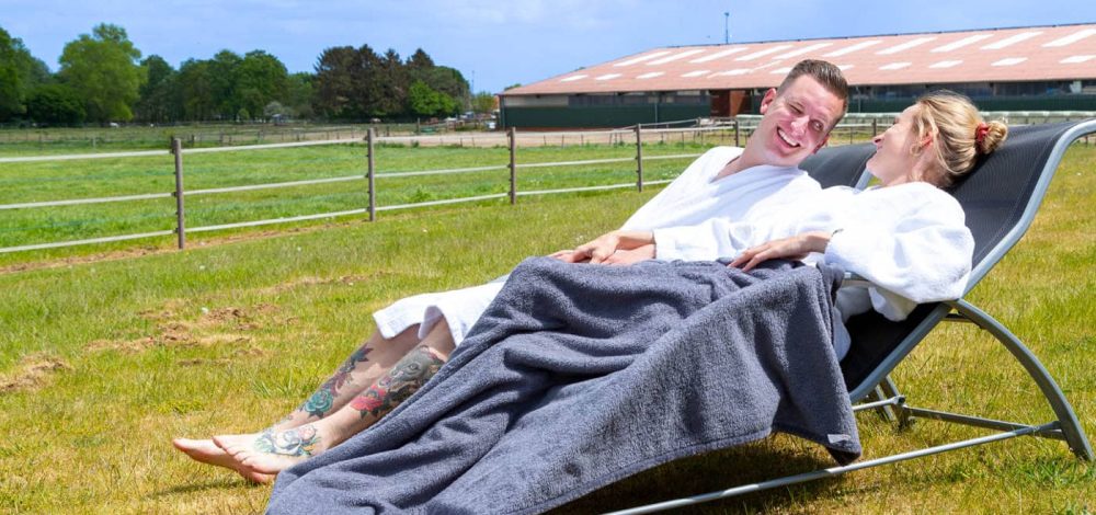 Familienhotel Gut Landegge im Emsland: Wellness als Paar. Mama und Papa liegen auf zwei liegen auf der Wiese, im Hintergrund der Reitstall mit Sandplatz.