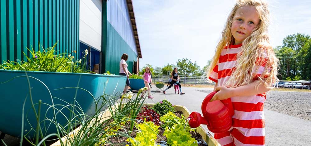Familienhotel Gut Landegge im Emsland: Kind gießt mit roter Gießkanne Salat im Hochbeet.